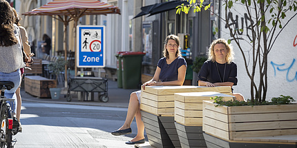 Zwei Frauen sitzen auf Holzbänken, daneben eine Frau am Fahrrad und ein Verkehrsschild.