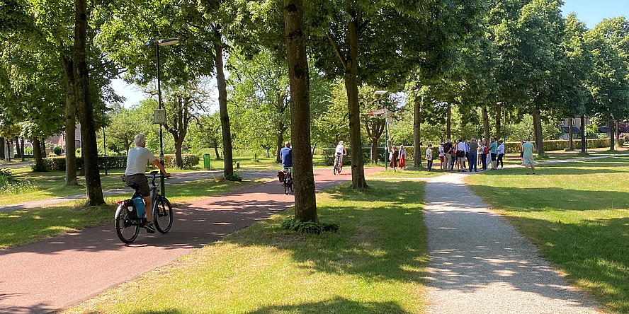 Parkanlage mit grünen Bäumen, Fahrradwegen mit Fahrradfahrern und Gehwegen mit einer Personengruppe im Hintergrund