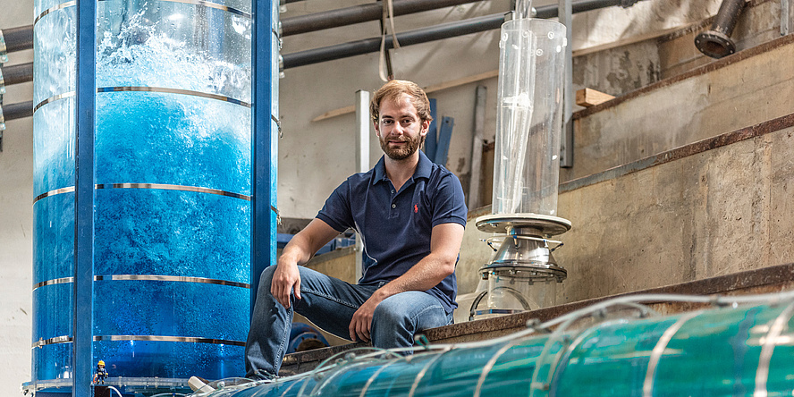 Ein junger Mann mit Bart in Jeans und blauem T-Shirt sitzt im Wasserbaulabor und ist umgeben von Versuchanordnungen in Form von wassergefüllten Plexiglas-Röhren