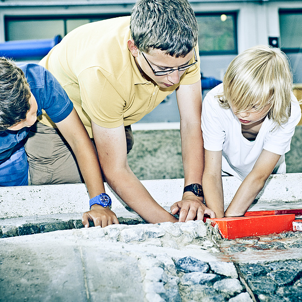 Zwei Schüler mit einem Studenten vor einem Modell mit Steinen. Bildquelle: Lunghammer – TU Graz