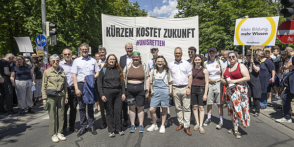 Zahlreiche Menschen stehen auf einer Straße und blicken in die Kamera. Hinter ihnen ist ein Banner mit der Aufschrift: "Kürzen kostet Zukunft! #unisretten"