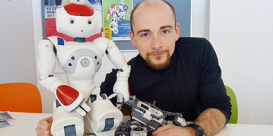[Translate to Englisch:] Martin Kandlhofer mit drei Robotern: einem humanoiden weißen Nao, einem Legoroboter und einem Würfelroboter.