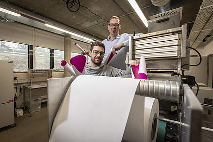 Two men stand behind a large roll of paper, holding up paper printed on one side.