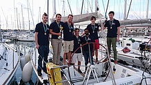 Sieben junge Männer auf einem Segelboot mit Medaillen und Pokal.