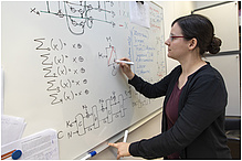 A woman is writing on a white board, several formulas and sketches can be seen.