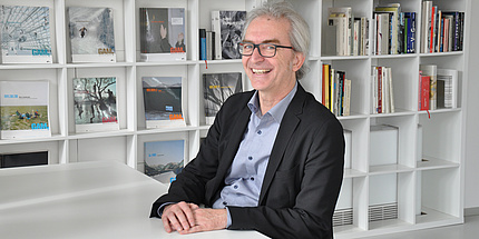 Grey-haired man at a desk in front of a white bookshelf.