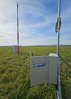 An antenna pole stands on a meadow and is connected by a cable to a metal box, which hangs from a smaller pole a few metres away.