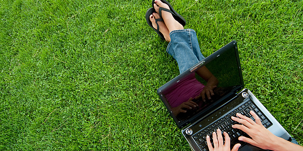 Eine Frau sitzt auf einer grünen Wiese und arbeitet mit dem aufgeklappten Laptop auf ihrem Schoß.