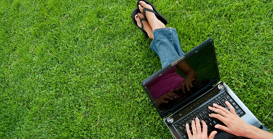 Eine Frau sitzt auf einer grünen Wiese und arbeitet mit dem aufgeklappten Laptop auf ihrem Schoß.