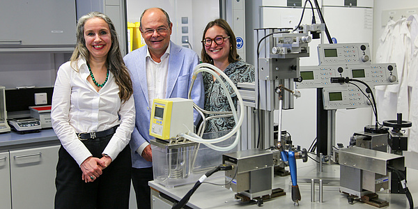 Three people, two women and a man, are standing in a laboratory and smile at the camera. The man is standing in the middle.