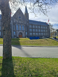 Historisches graues Steingebäude mit Spitzdach und Türmen, davor große blaue NTNU-Buchstaben auf einer Rasenfläche mit norwegischen Flaggen und kahlen Bäumen im Vordergrund.
