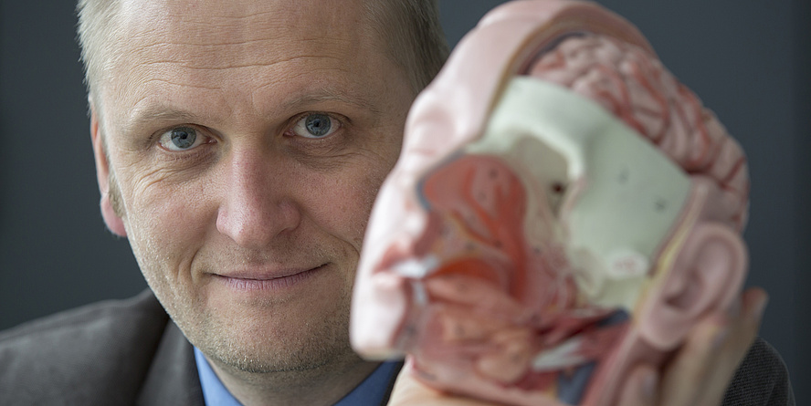 Ein blonder Mann, der eine Plastikabbildung eines Schädels in der Hand hält.