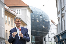 Mann mit grauen Haaren und dunklem Anzug steht vor dem Kunsthaus in Graz und schleckt ein Eis. 
