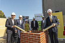 5 Männer und eine Frau stehen auf einer Baustelle - sie tragen weiße Bauhelme. Im Vordergrund sind Ziegelsteine.