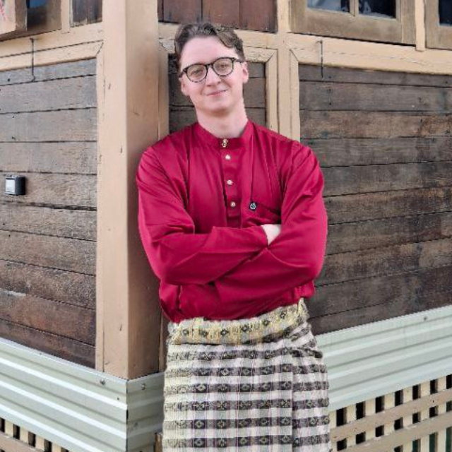 Ein junger Mann steht in einer traditionellen rot-beigen Robe mit verschränkten Armen vor einem Holzhaus. 