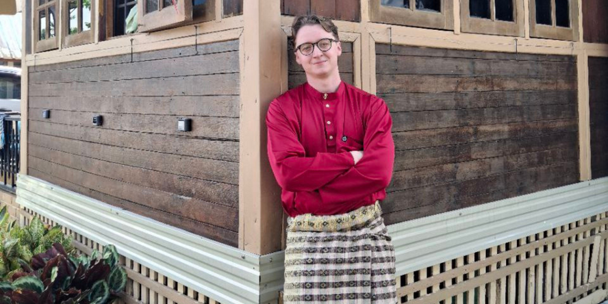Ein junger Mann steht in einer traditionellen rot-beigen Robe mit verschränkten Armen vor einem Holzhaus. 