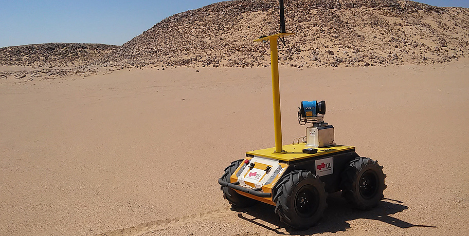Brauner Wüstensand. Steine im Hintergrund. Vorne steht ein Roboter - er ist gelb, geformt wie eine Kiste und hat eine lange Antenne.