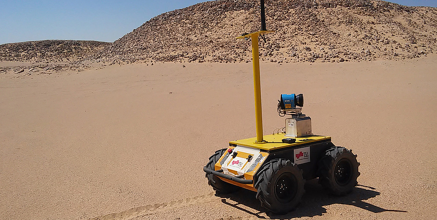 Brown desert sand. Stones in the background. At the front there is a robot - it's yellow, shaped like a box, and has a long antenna.