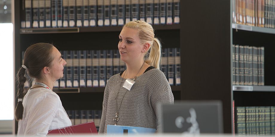 Zwei Frauen stehen vor einem Bücherregal in einer Bibliothek und unterhalten sich