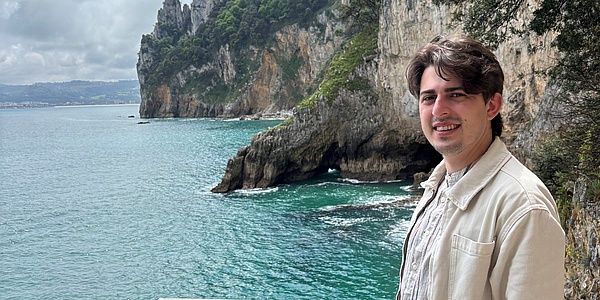 A young man smiles at the camera, with rugged but green rock cliffs rising out of the turquoise blue water in the background.