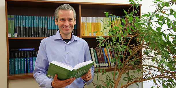 A man in a blue shirt stands smiling in front of a bookshelf. He is holding an open book in his hands. Next to him is a houseplant.