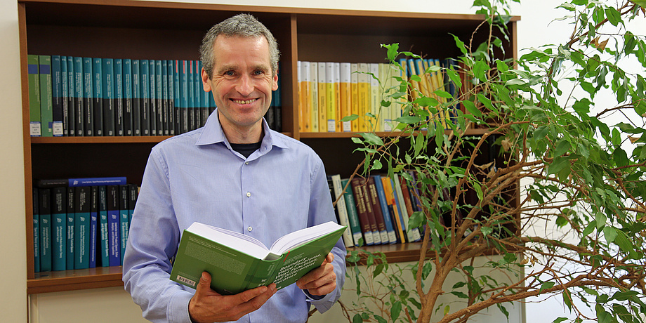 A man in a blue shirt stands smiling in front of a bookshelf. He is holding an open book in his hands. Next to him is a houseplant.