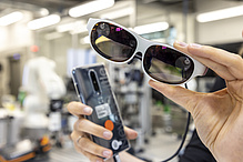 Eine Hand hält eine Datenbrille in die Kamera, im Hintergrund ist ein Smartphone zu sehen. 