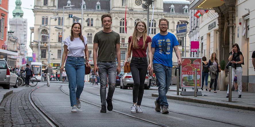 Zwei Studentinnen und zwei Studenten gehen vor dem Hintergrund des Grazer Hauptplatzes auf den Straßenbahnschienen dem Fotografen entgegen.