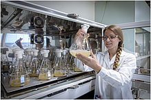 A woman in a lab coat swings a glass flask with light liquid.