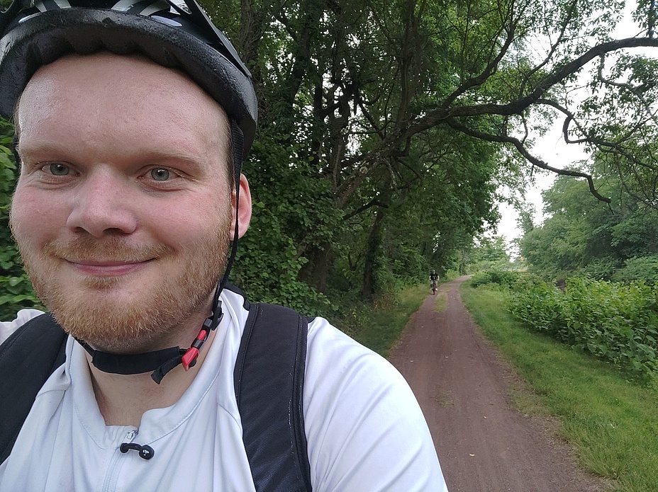 Mann mit weißen Fahrradhelm blickt in die Kamera, im Hintergrund sieht man Fahrradwege.