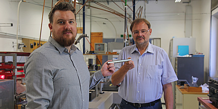Peter Pichler und Gernot Pottlacher stehen in einem Labor und halten einen Eisenstab in der Hand. 