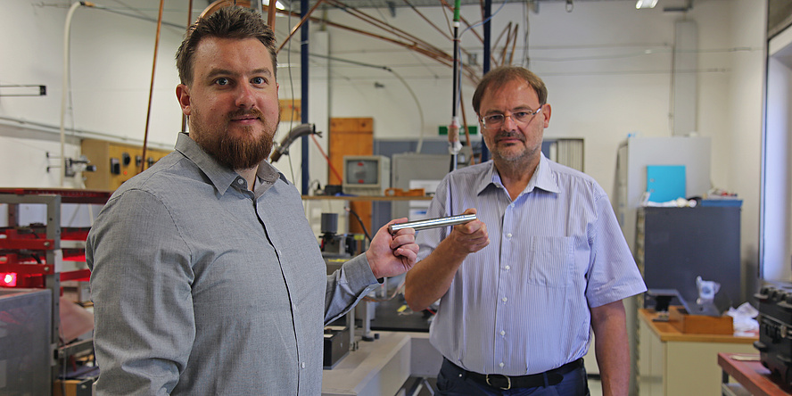 Peter Pichler und Gernot Pottlacher stehen in einem Labor und halten einen Eisenstab in der Hand. 