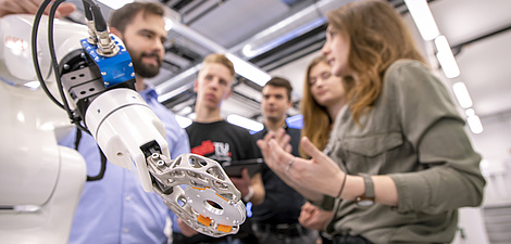 Ein Maschinenarm im Vordergrund, dahinter stehen Menschen, die miteinander sprechen