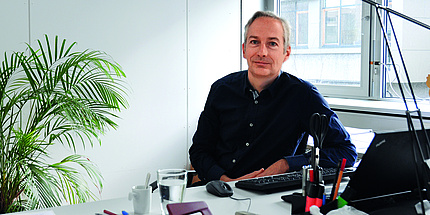 A man with blue eyes, greying hair and dark shirt sits at his desk between a big window and an indoor palm.