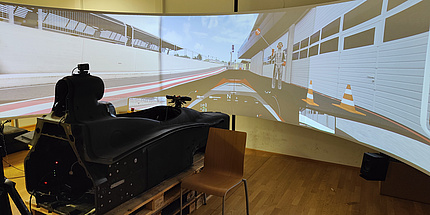 A cockpit of a formula racing car stands in front of a screen on which the pit lane of a race track is shown in computer graphics.