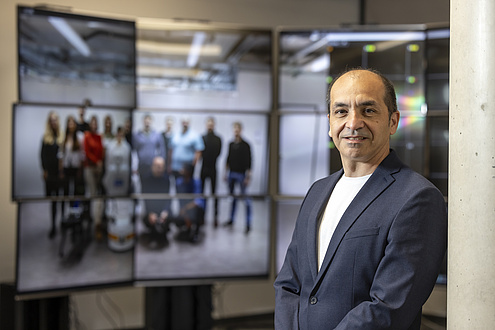 A man stands in front of a row of monitors and smiles at the camera