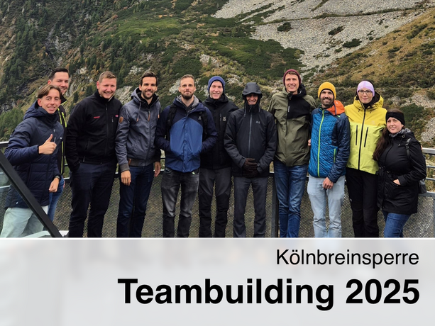 Eine Gruppe von zehn Personen in Outdoorjacken posiert lächelnd vor der Kölnbreinsperre in den Bergen, mit felsigen Hängen und grünen Almflächen im Hintergrund; Text: „Kölnbreinsperre Teambuilding 2025“.  
