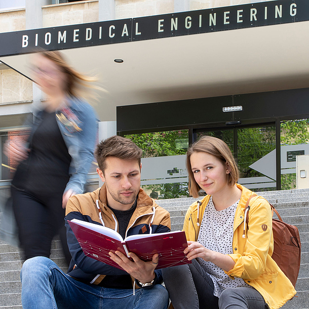 Zwei Studierende sitzen auf Stufen vor einem Gebäude mit der Aufschrift "Biomedical Engineering".