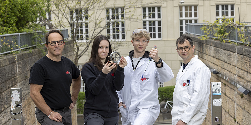 Three men and a woman are standing on a staircase. Two of the men are wearing white lab coats, while the woman and the other man are wearing black T-shirts. The coats and T-shirts are printed with the TU Graz logo. 