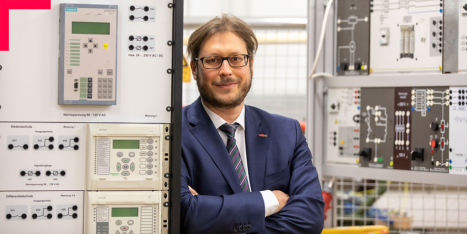 Man with beard and glasses in an electrical engineering laboratory, leaning against a piece of equipment