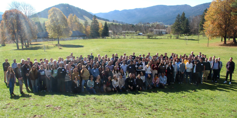 Gruppenfoto von rund 200 Personen auf einer großen Wiese mit Wald und Bergen im Hintergrund.