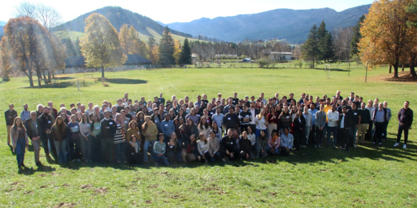 Gruppenfoto von rund 200 Personen auf einer großen Wiese mit Wald und Bergen im Hintergrund.