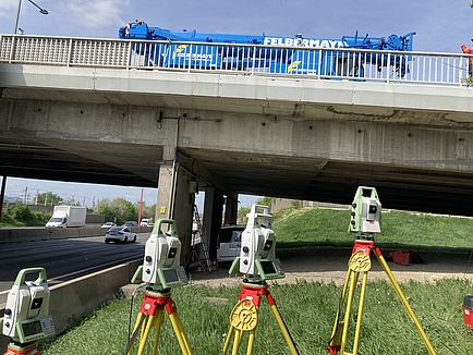 Ein blauer Kranwagen steht auf einer Brücke. Davor sind auf einer Wiese einige Messgeräte zu sehen.
