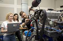 Ein Mann schraubt an einer Maschine, zwei Frauen beobachten prüfend.