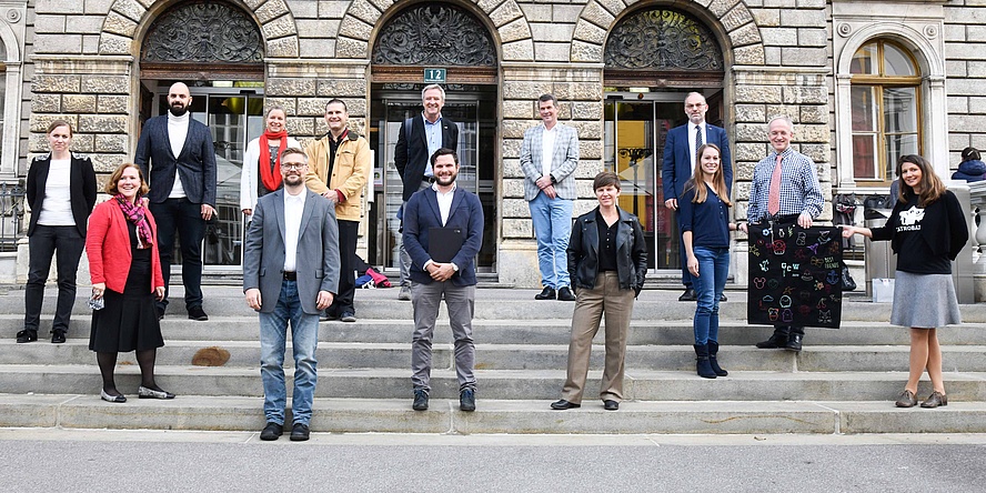 14 Menschen unterschiedlichen Alters am Stiegenaufgang zu einem historischen Gebäude.