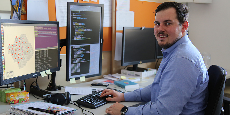 A young man is sitting at a desk. He wears a blue shirt, short brown hair and a short beard. His right hand touches the keybord. On the computerscreen there is a grey grafic with red elements.