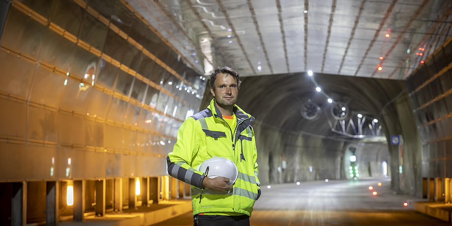 Mann mit Schutzjacke und Helm unter dem Arm steht vor einer Tunneleinfahrt.