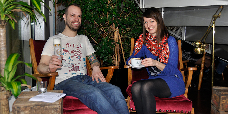 A man with a beard, a tattoo on his arm and a beer in his hand as well as a woman with medium-length brown hair, purple long-sleeved sweater and red cloth and a cup of coffee in hand sit on comfortable chairs covered with red velvet armchairs, green plants behind them.