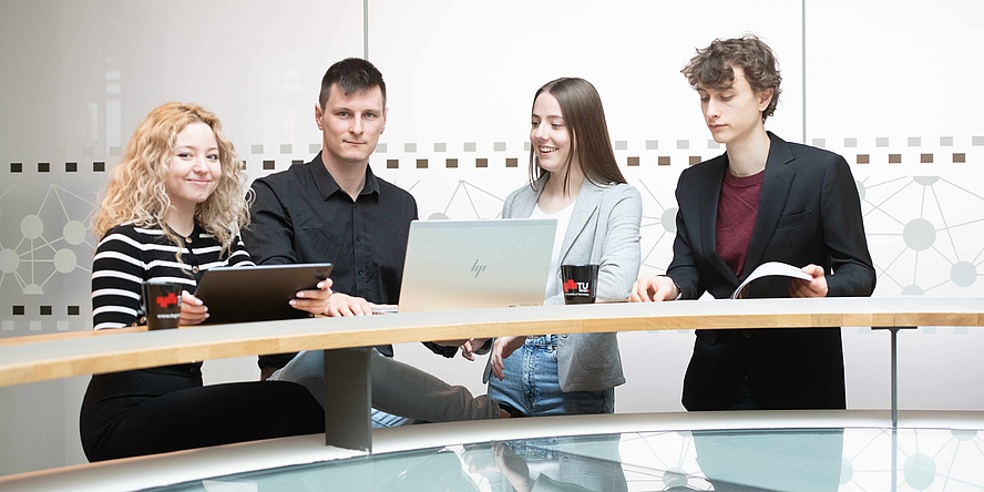 Zwei junge Frauen und zwei junge Männer mit Laptop und Tablet, geschäftsmäßig gekleidet, stehen an einem langen Stehtisch.