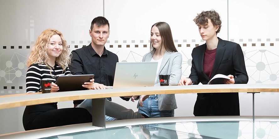 Two young women and two young men with laptop and tablet, dressed business-like, stand at a long bar table.